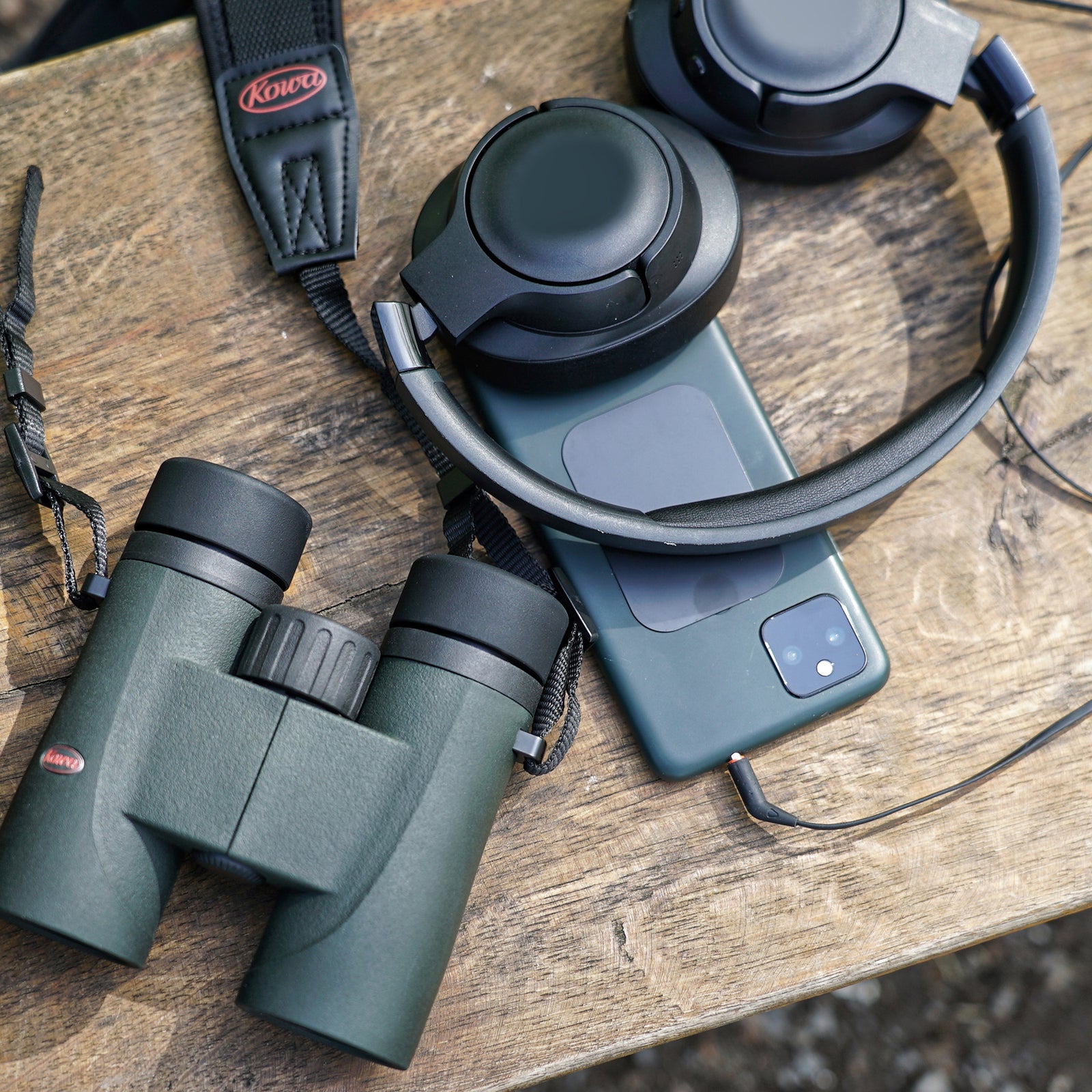 Kowa SV III binoculars beside a phone and headphones on an outdoor table, showing their place in an everyday travel kit